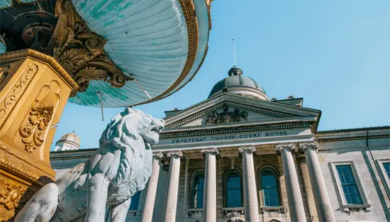 Looking up at the historic Frontenac County Courthouse in Kingston, with a bronze lion statue and ornate water fountain in the foreground.